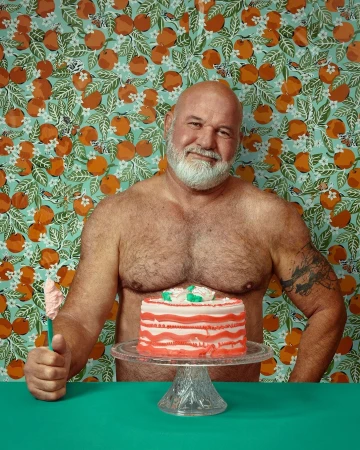 Man poses topless with a cake on a stand in front of a patterned wallpaper; he has a white beard and a tattoo on his arm.